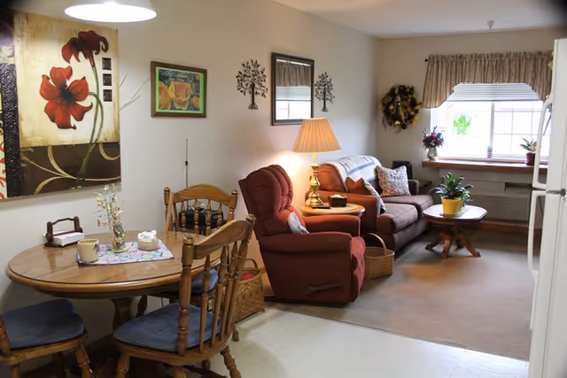 A cozy living room and dining area in a senior living facility with a round wooden dining table and four chairs, a red recliner chair, a brown sofa, a wooden coffee table with a potted plant, a side table with a lamp, wall decorations including paintings and metal tree art, and a window with curtains letting in natural light.