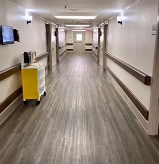 Long, well-lit nursing home hallway with handrails, closed doors, and a small yellow cart on the left.