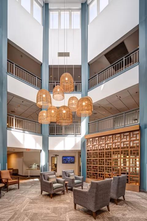 A spacious, multi-level common area with tall ceilings and large windows letting in natural light. The room features a cluster of hanging wicker pendant lights, several gray upholstered armchairs arranged in small groups, and a decorative wooden lattice partition. The upper levels have railings overlooking the area below.