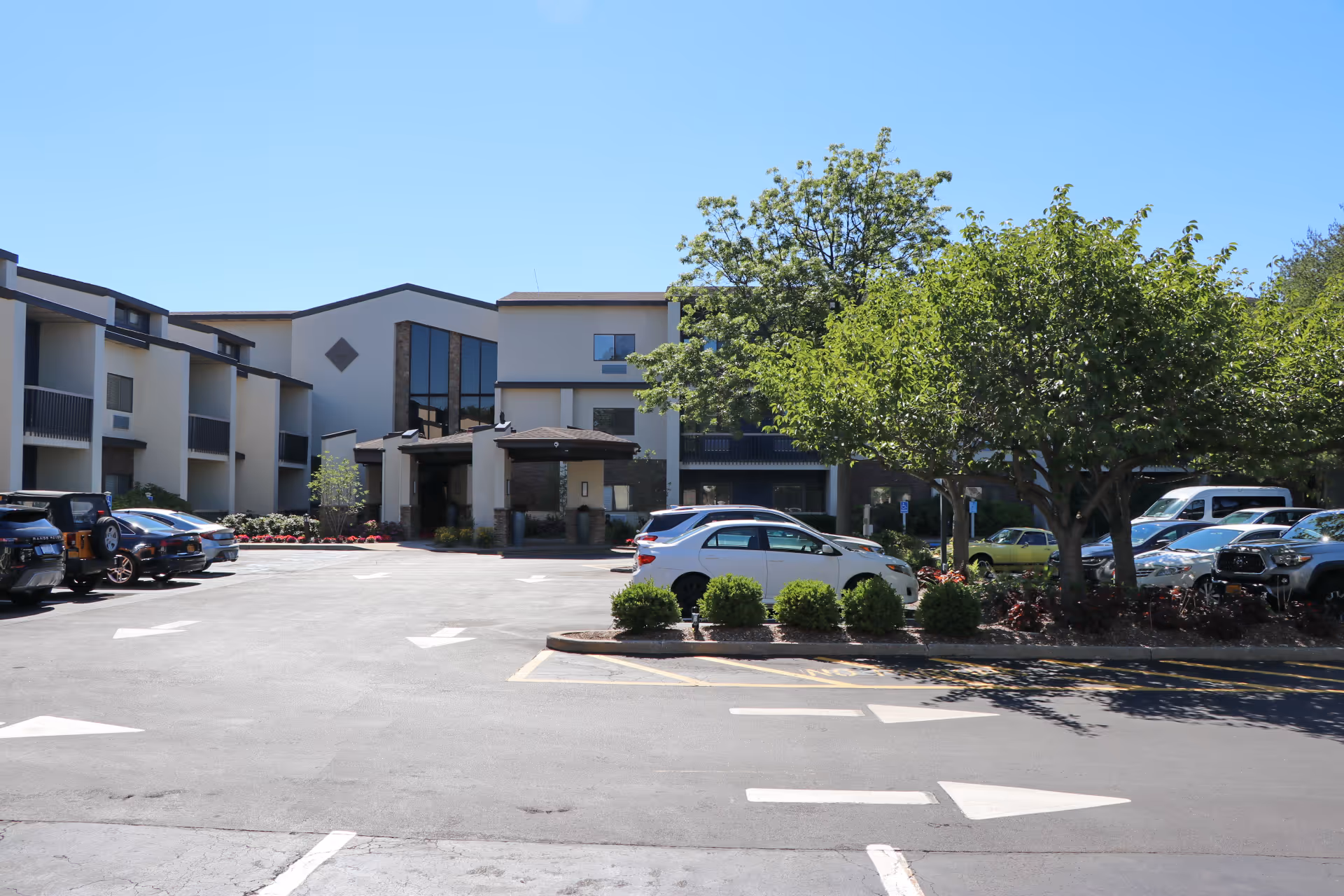 Exterior view of The Residences at Plainview by Chelsea Senior Living showing a multi-story building with balconies, a parking lot with several parked cars, and trees providing shade under a clear blue sky.