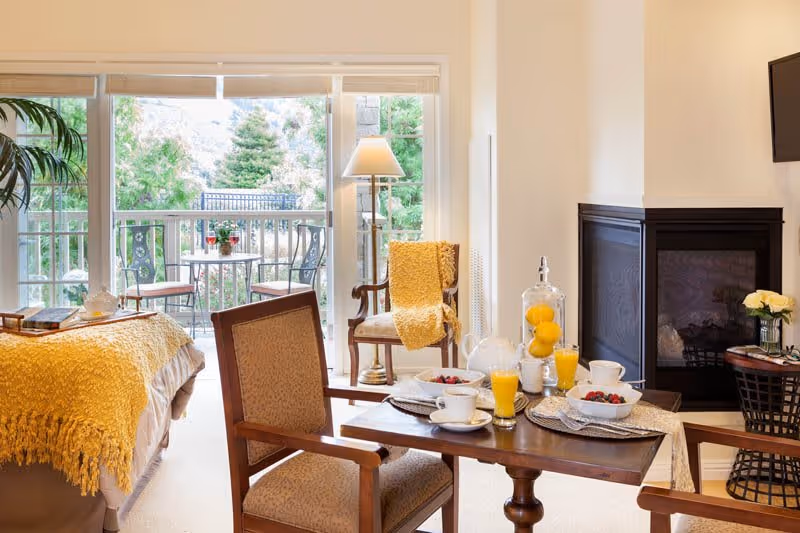 A cozy living area in a senior living facility with a small wooden dining table set for breakfast, including glasses of orange juice, cups, and bowls of fruit. There are two upholstered chairs around the table. A fireplace is visible on the right side with a small table holding a vase of flowers. A comfortable chair with a yellow throw blanket is next to a floor lamp. Large sliding glass doors open to a balcony with a small table and two chairs, overlooking greenery and trees.
