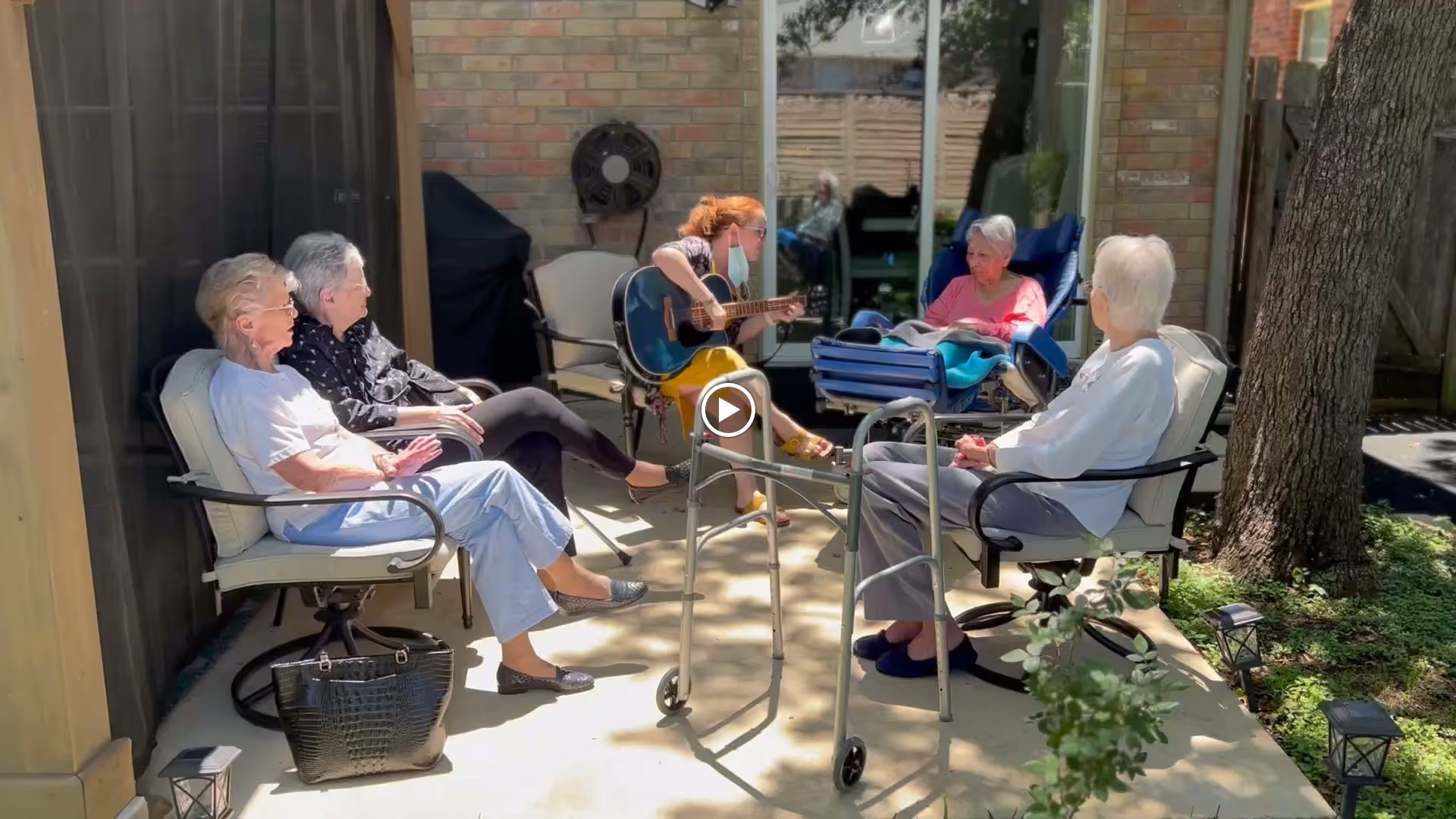 Five residents seated on a shaded outdoor patio while a person plays an acoustic guitar.