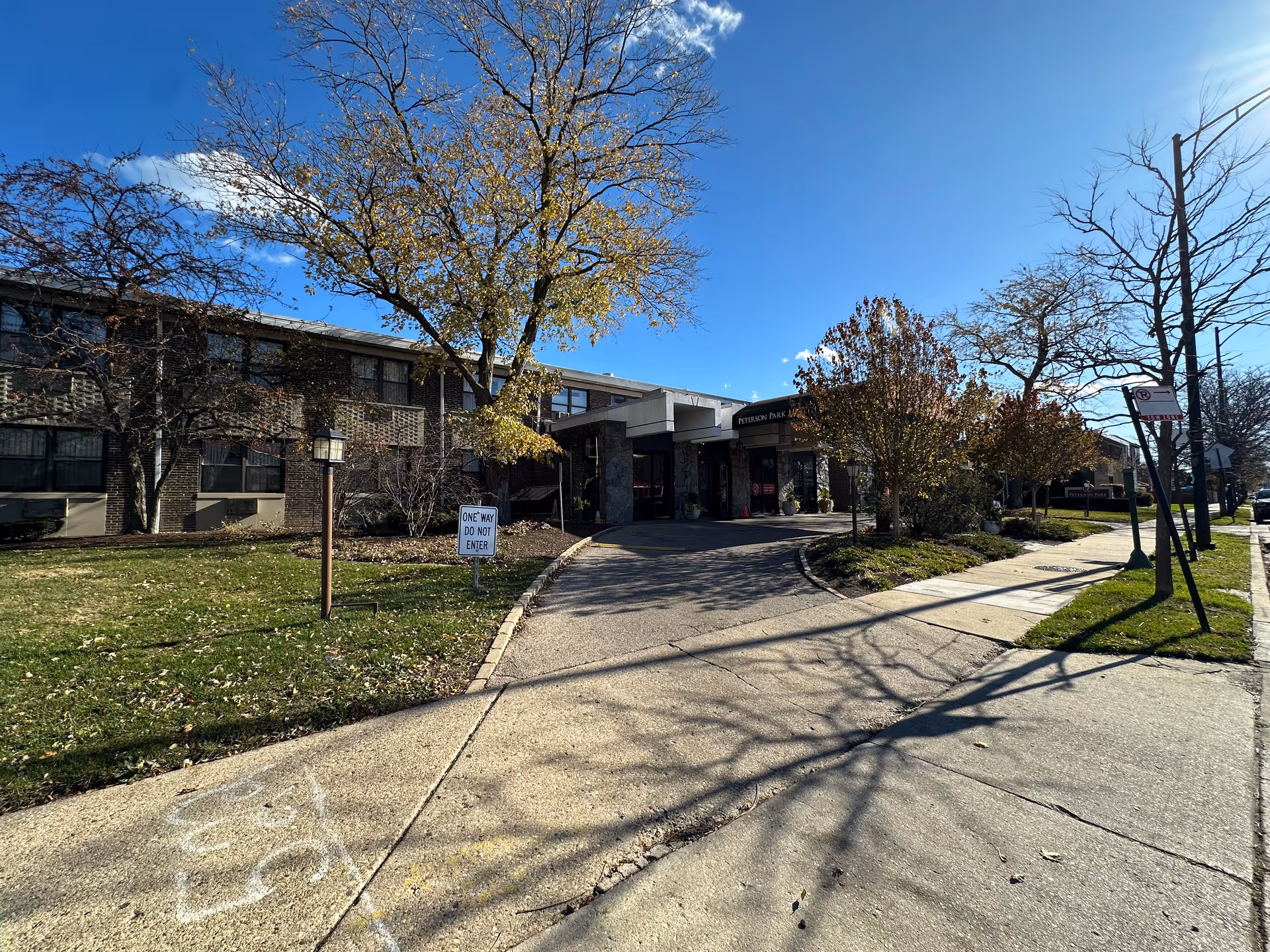 Exterior view of Peterson Park Health Care Center on a clear day with blue sky. The building is two stories tall with brick walls and multiple windows. There is a driveway leading to the main entrance, surrounded by trees with autumn foliage and a grassy area. A sign near the driveway reads 'ONE WAY DO NOT ENTER'.