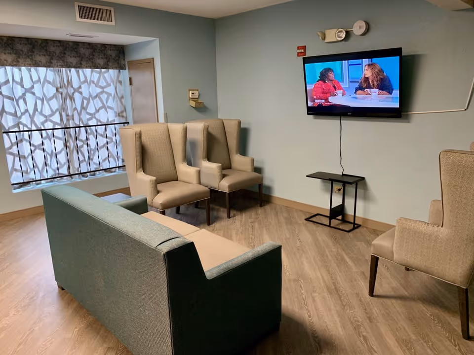 Communal sitting area with a sofa and several wingback chairs facing a wall-mounted TV in a senior living facility.