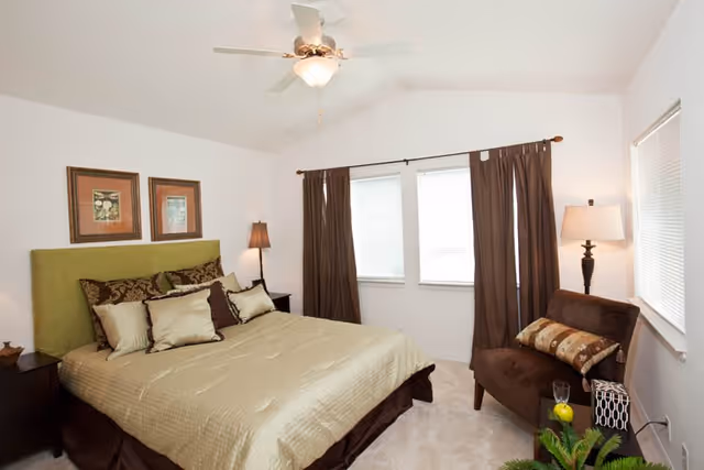 A bright bedroom with a large bed featuring a green headboard and multiple pillows. There are two framed pictures on the wall above the bed. The room has two windows with brown curtains, a ceiling fan with a light, a brown armchair with a decorative pillow, a floor lamp, and a small plant on the floor.