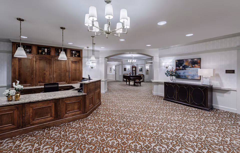A senior living facility lobby with a wooden reception desk, patterned carpet, chandeliers, and a seating area beyond.