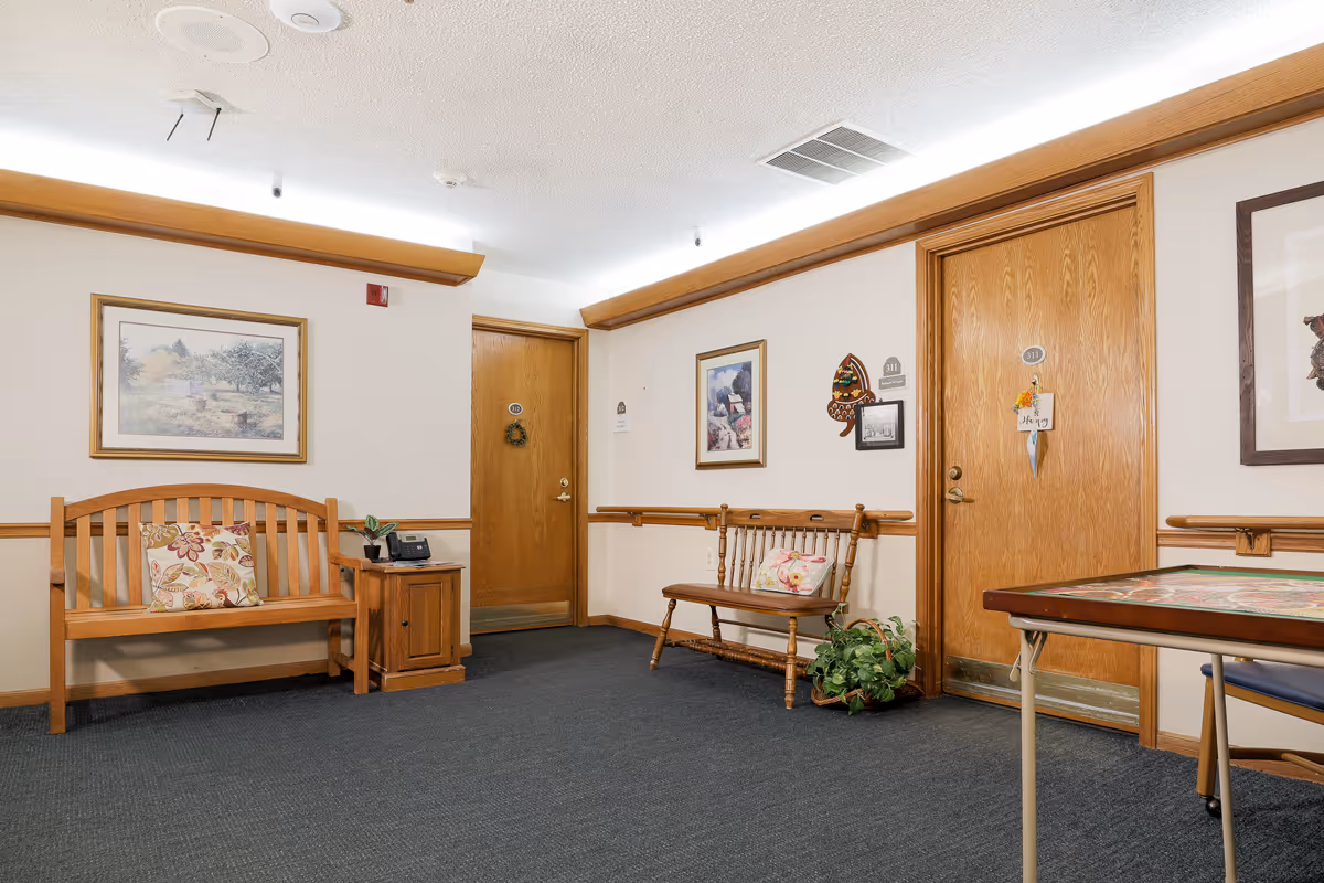 Carpeted interior hallway of an assisted living facility with wooden benches, framed artwork, and apartment doors.