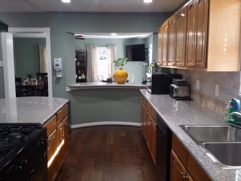 A modern kitchen with wooden cabinets, granite countertops, a double sink, a stove, and various small appliances. The kitchen opens to a living area with a window, curtains, a wall-mounted TV, and a yellow vase with a plant on the counter.