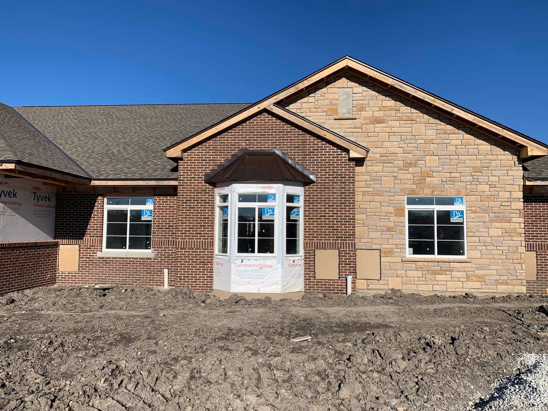 Exterior view of a building under construction with a combination of brick and stone facade, multiple windows, and a bay window in the center. The ground in front is bare dirt with construction marks, and the sky is clear and blue.
