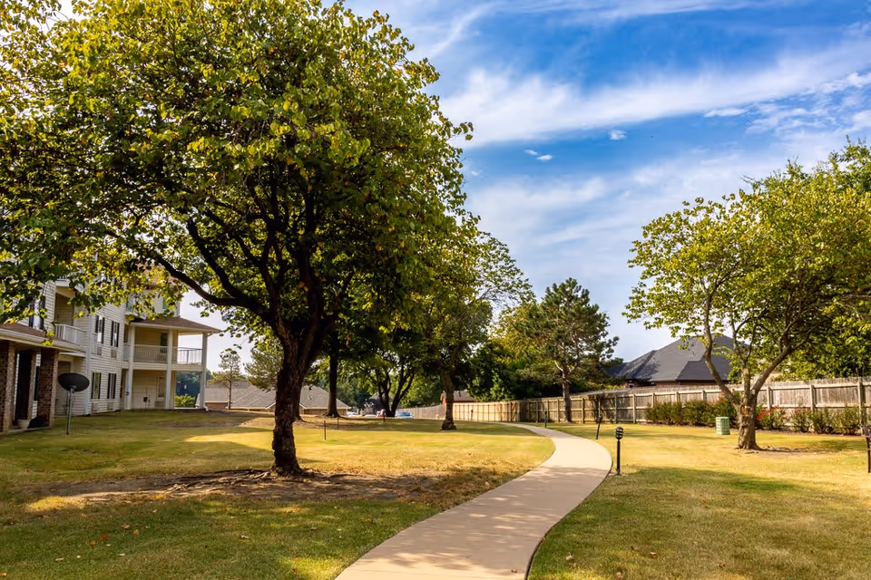 A winding concrete pathway through a grassy outdoor area with several trees and a wooden fence in the background. On the left side, there is a multi-story residential building with balconies. The sky is partly cloudy with blue patches.