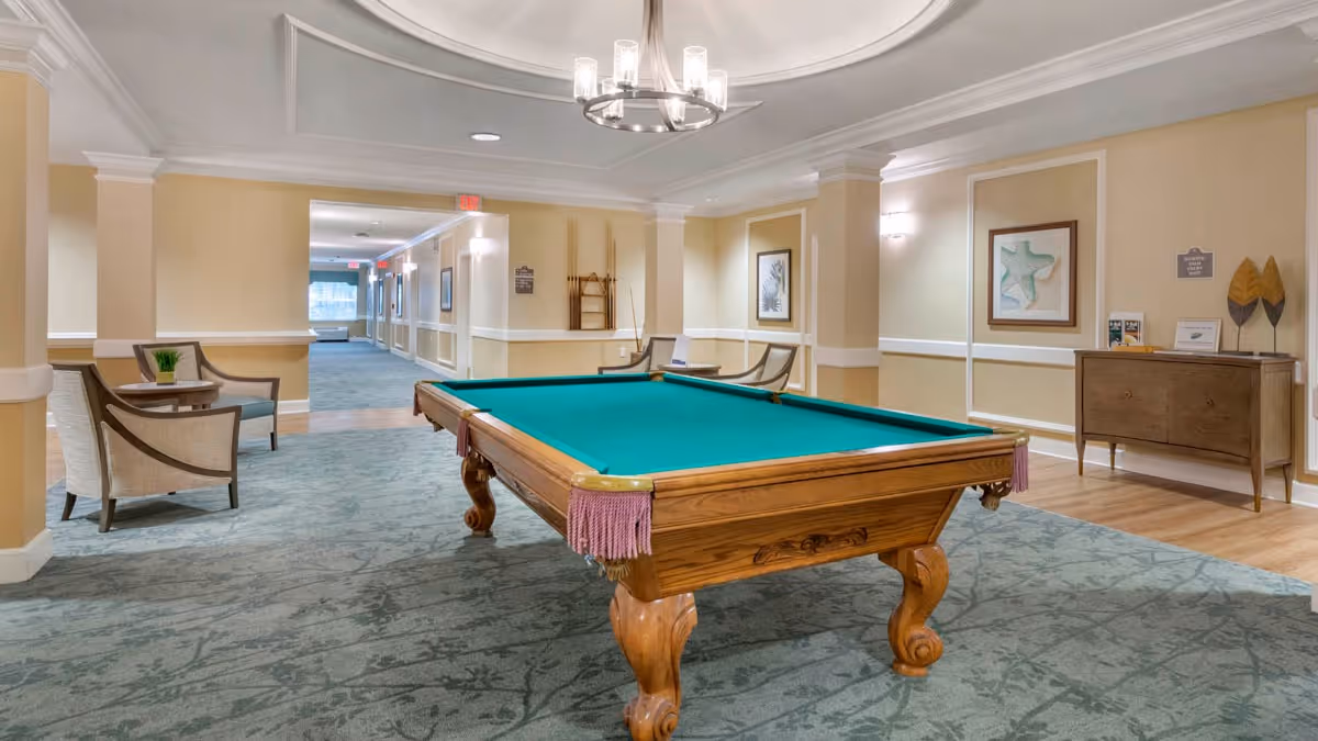 A spacious indoor common area with a green felt pool table in the center, surrounded by beige walls with white trim. There are two chairs with a small table on the left side and a wooden sideboard with decorative items on the right. The ceiling has a modern chandelier, and the floor is carpeted with a patterned design. A hallway extends into the background with additional seating and framed artwork on the walls.