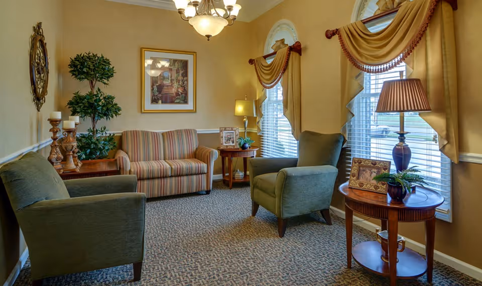 A cozy sitting area in a senior living facility with two green armchairs and a striped loveseat arranged around two wooden side tables. The tables hold lamps, framed photos, and decorative plants. The room has beige walls, carpeted floor, two windows with blinds and gold drapes, and a framed painting on the wall.