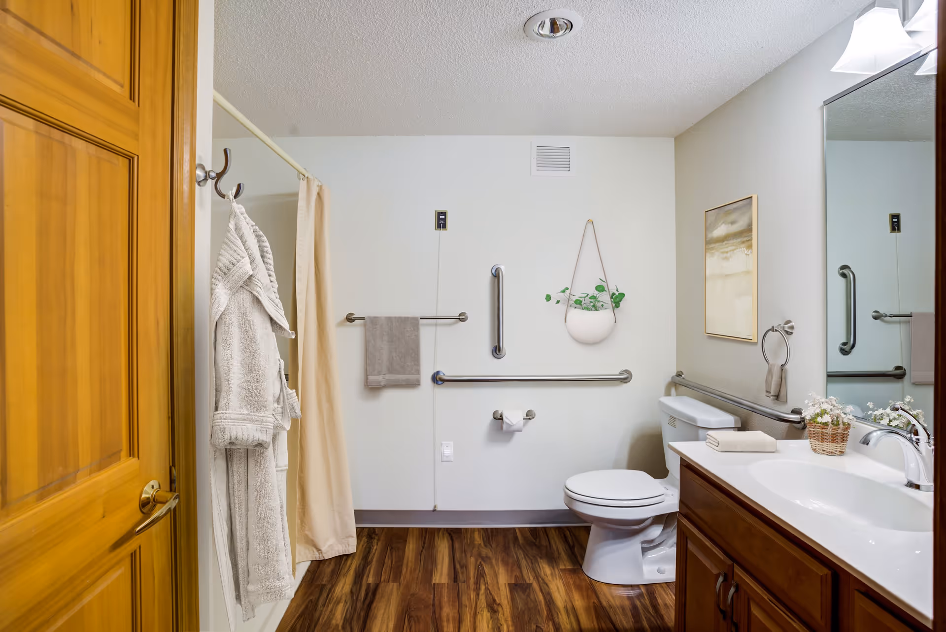 A clean, accessible bathroom with wooden flooring, a white toilet, a wooden vanity with a white sink, a large mirror, and a hanging plant on the wall. There are grab bars around the toilet and a beige shower curtain with a bathrobe hanging on a hook on the door.