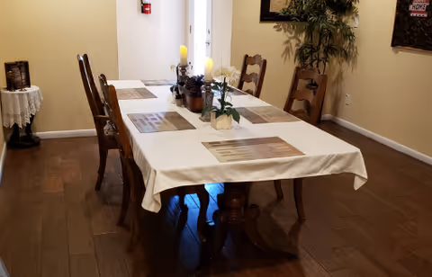 A rectangular dining table covered with a white tablecloth, placemats, candles, and a floral centerpiece surrounded by wooden chairs in a warm interior.