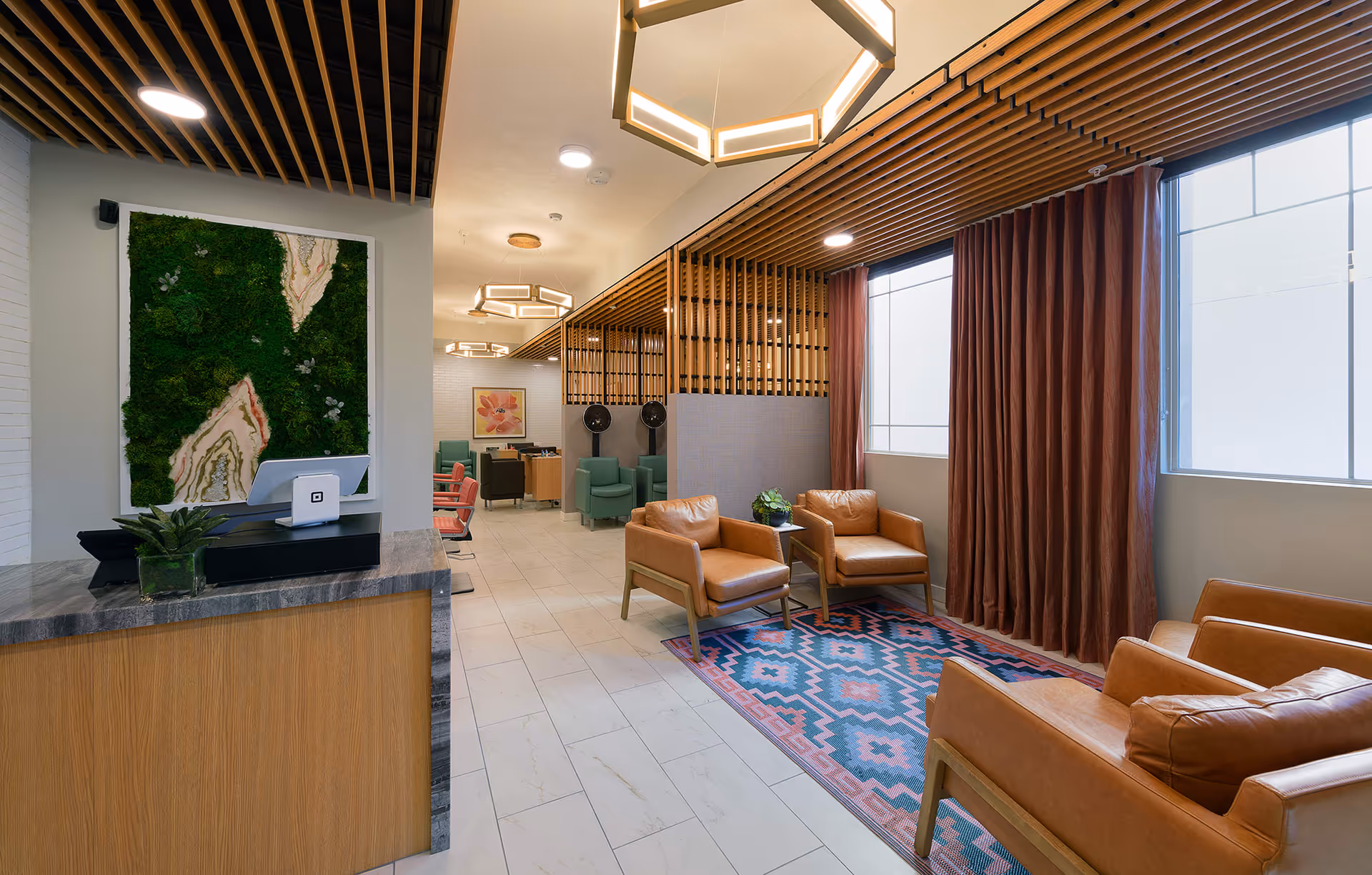 A modern senior living facility lounge area with tan leather armchairs arranged around a colorful patterned rug. The space features large windows with brown curtains, wooden slatted ceiling accents, and geometric pendant lights. A reception desk with a small plant and a green moss wall art piece is visible on the left. In the background, there are additional seating areas with green and pink chairs and a floral painting on the wall.
