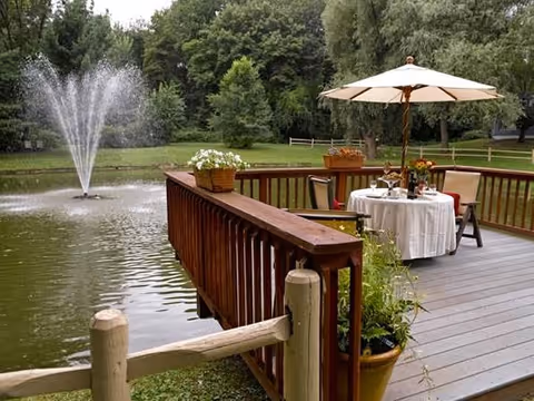 A wooden deck overlooking a pond with a water fountain. The deck has a round table covered with a white tablecloth, set for dining with chairs and an umbrella. There are potted plants on the deck railing and a wooden fence surrounding the pond. Trees and greenery are visible in the background.