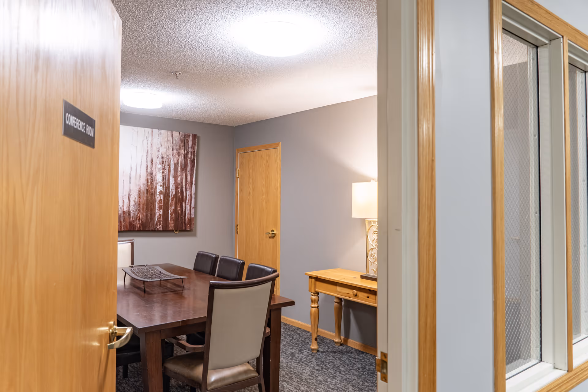 View into a conference room with a wooden table surrounded by chairs. A decorative metal tray is on the table. The room has gray walls, a wooden door, a wooden side table with a lamp, and a large artwork featuring abstract tree trunks on the wall.