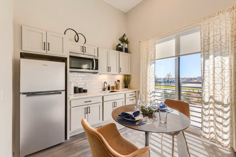 A bright kitchen area with white cabinets, a stainless steel refrigerator, microwave, and sink. A round dining table with two brown chairs is set with plates, glasses, and napkins. Large windows with sheer patterned curtains let in natural light and offer a view of the outdoors.