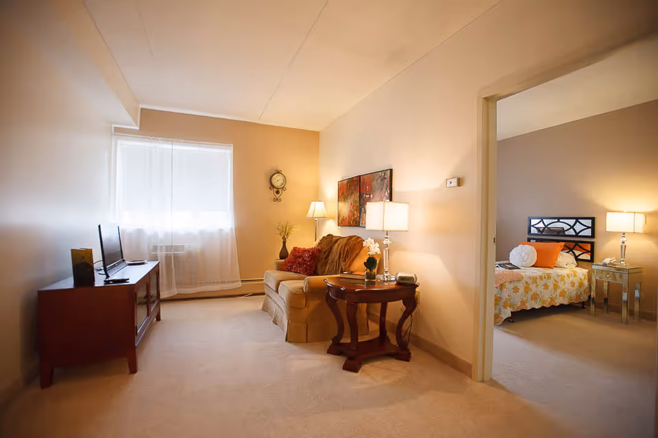 A cozy living room area with a beige armchair, a wooden side table with a lamp and a small plant, and a TV on a wooden stand. The room has beige walls and carpeted floor. Through an open doorway, a bedroom is visible with a bed covered in a floral bedspread, a round white pillow, an orange pillow, a bedside table with a lamp, and a beige wall.