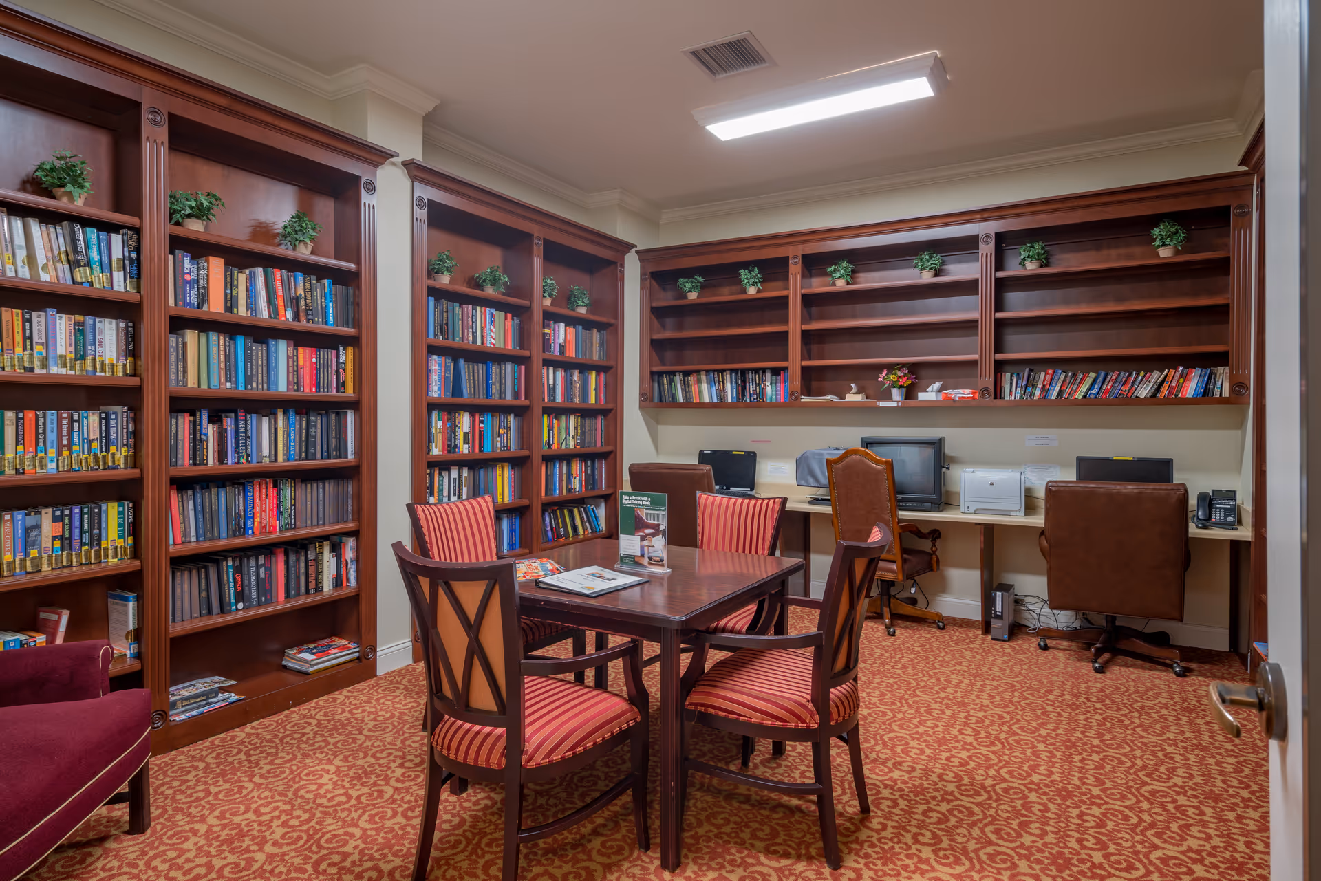 A cozy library room with wooden bookshelves filled with books and small potted plants on top. There is a wooden table with four red and brown cushioned chairs in the center of the room. Along the back wall, there is a desk with three computer workstations, a printer, and office chairs. The room has a patterned carpet and a bright rectangular ceiling light.