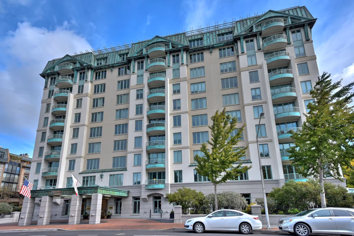 Front exterior of The Stratford, a multi-story residential building with green balconies and roof, an entrance canopy with flags, trees, and parked cars.