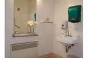 Small tiled bathroom with a wall-mounted sink, mirror, soap and paper-towel dispensers, grab bar and a vase of white flowers.