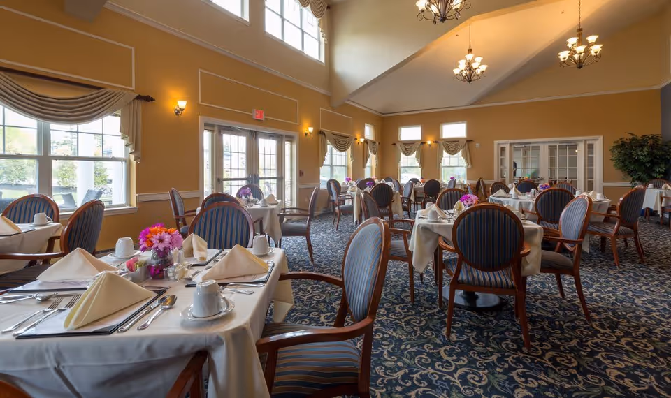 A spacious dining room with multiple round tables covered with white tablecloths, each set with folded napkins, cups, and silverware. The room features large windows with draped curtains, high ceilings with chandeliers, and patterned carpet flooring. There are floral centerpieces on the tables and a glass door leading outside.