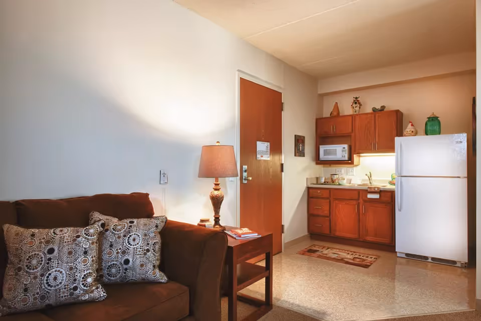 Cozy studio-style living area with a brown sofa and patterned pillows and a side table lamp adjacent to a compact kitchenette with wooden cabinets and a white refrigerator.