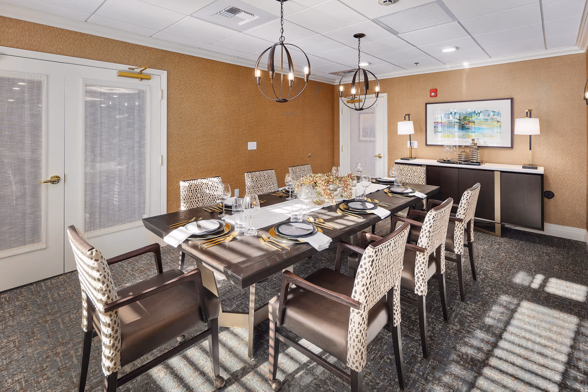 A well-lit dining room with a long dark wooden table set for eight people. The table is adorned with plates, glasses, gold-colored cutlery, white napkins, and a floral centerpiece. The room has patterned chairs with dark seats and light backs, two modern spherical chandeliers hanging from the ceiling, and a sideboard with two lamps and a framed abstract painting on the wall. Sunlight streams through a window, casting shadows on the carpeted floor.