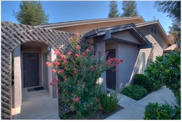 Exterior view of a senior cottage apartment at Arbor Manor Senior Cottage Apartments, featuring a beige building with a dark door, a wooden lattice archway, and vibrant pink flowering bushes along a concrete pathway surrounded by greenery.