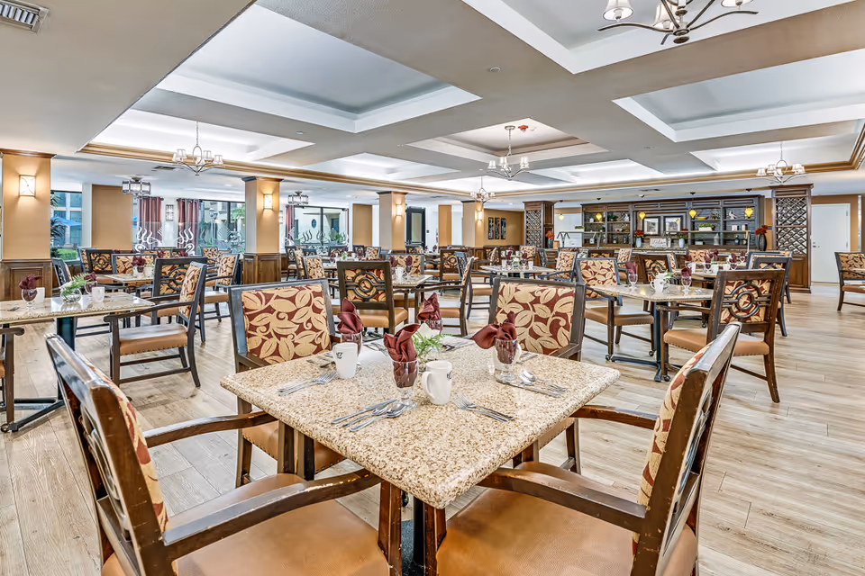 A spacious and well-lit dining room with multiple tables and chairs arranged neatly. Each table is set with utensils, napkins folded in glasses, and small decorative plants. The room features wooden flooring, beige walls with wooden paneling, and ceiling lights with chandeliers. Large windows allow natural light to enter, and there is a counter or bar area at the back with shelves and decorative items.
