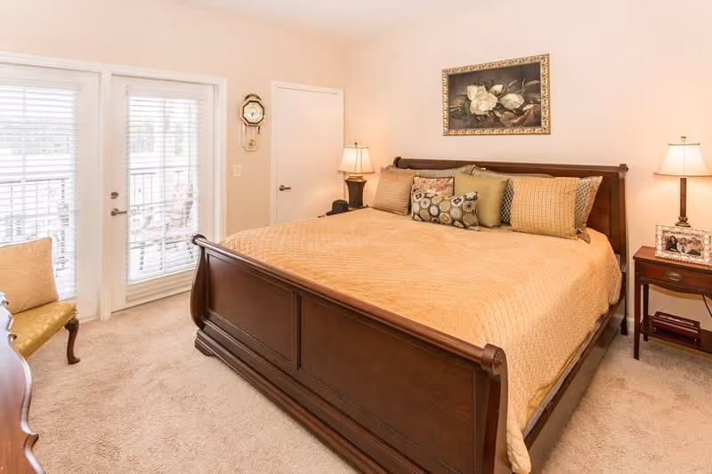 A bedroom with a large wooden bed covered with a beige quilt and multiple decorative pillows. There are two bedside tables with lamps on each side of the bed. A framed floral painting hangs on the wall above the bed. To the left, there are glass doors with blinds leading outside, and a small upholstered chair is positioned near the doors. The room has beige walls and carpeted flooring.