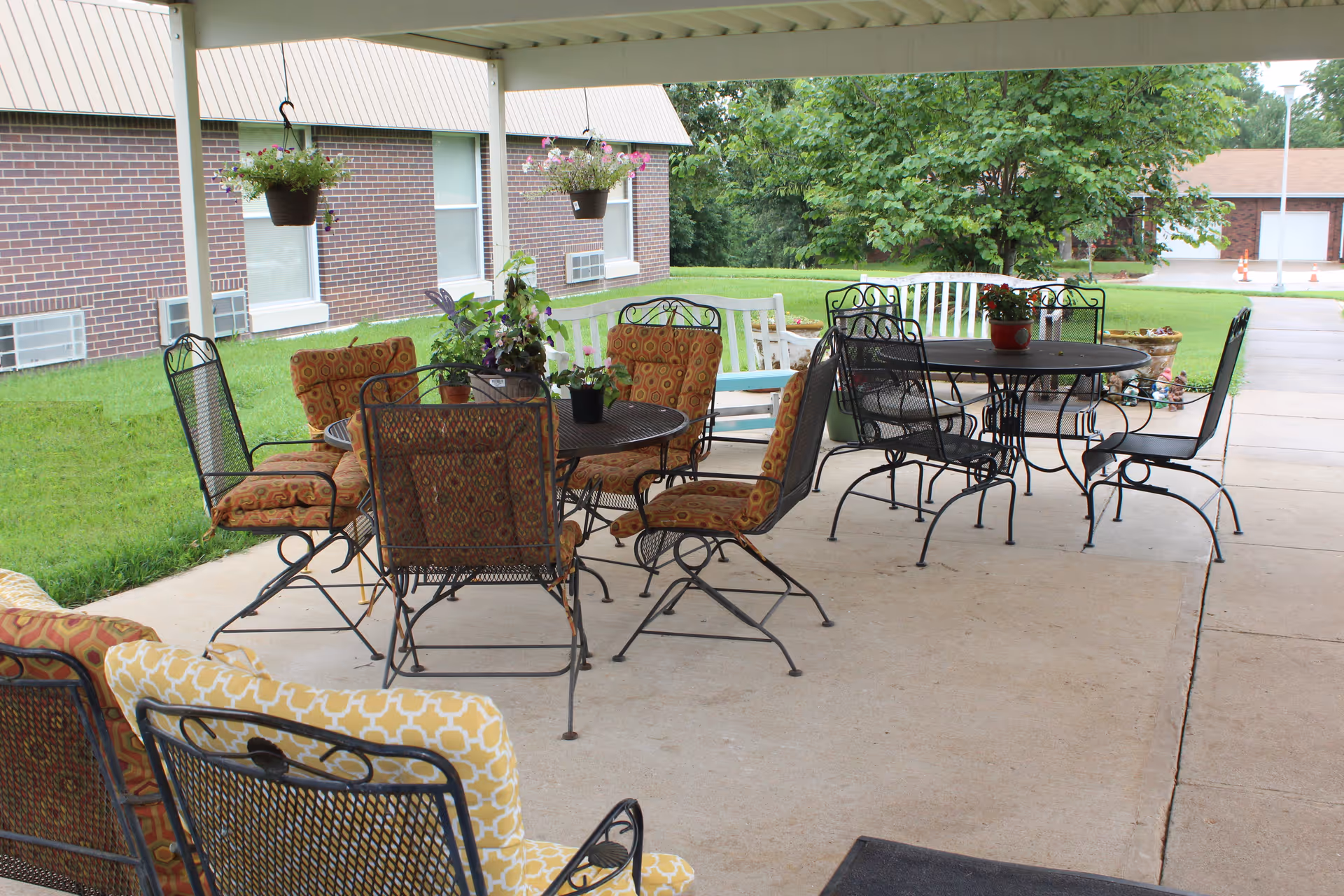 Covered outdoor patio with metal tables, cushioned chairs, hanging plants, and a grassy lawn by a brick building.