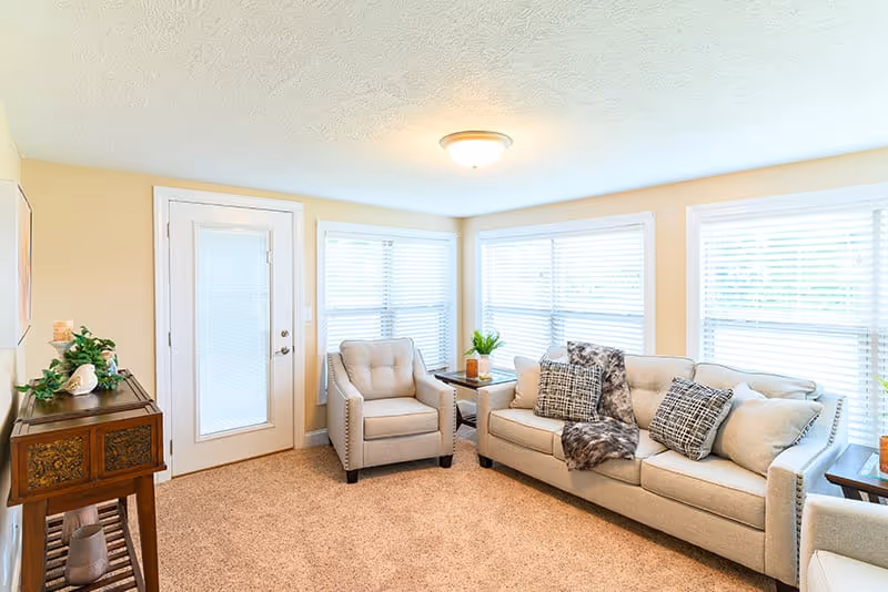 A bright living room with beige walls and carpet, featuring a beige sofa with patterned pillows and a throw blanket, a matching armchair, a wooden side table with a plant, and a wooden console table with decorative items. Large windows with white blinds allow natural light to fill the room.