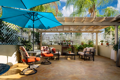 Outdoor patio area with cushioned chairs, a table, potted plants, and a large blue umbrella providing shade. The space is enclosed with lattice fencing and has a pergola overhead. Palm trees and a blue sky with clouds are visible in the background.