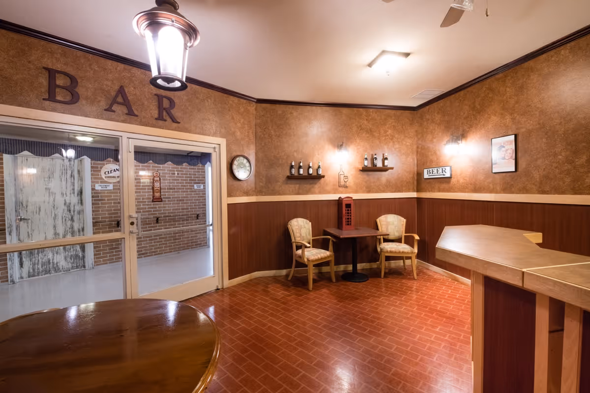 Cozy indoor bar/lounge area with a small wooden bar, two chairs and a table, shelves with bottles, and double glass doors beneath a 'BAR' sign.