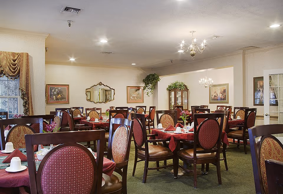 Dining room with multiple round tables set with red tablecloths and wooden chairs in a senior living facility.