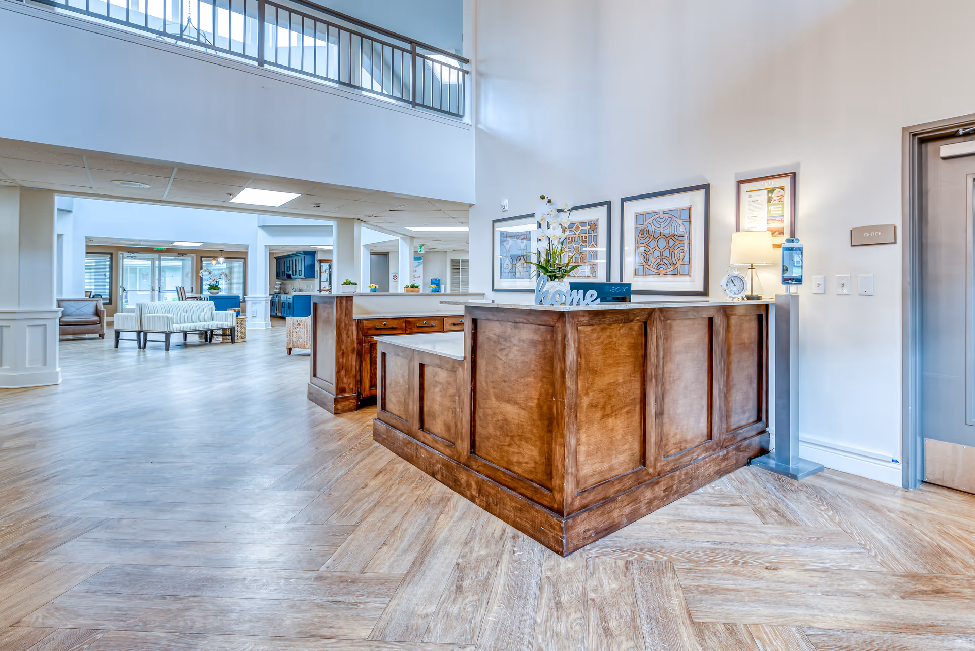 Bright and spacious senior living facility interior with a wooden reception desk in the foreground, decorated with a small plant and framed artwork on the wall behind. The room features light wood flooring, white walls, and a high ceiling with large windows allowing natural light. In the background, there are seating areas with sofas and chairs, and a kitchen area with blue cabinetry.