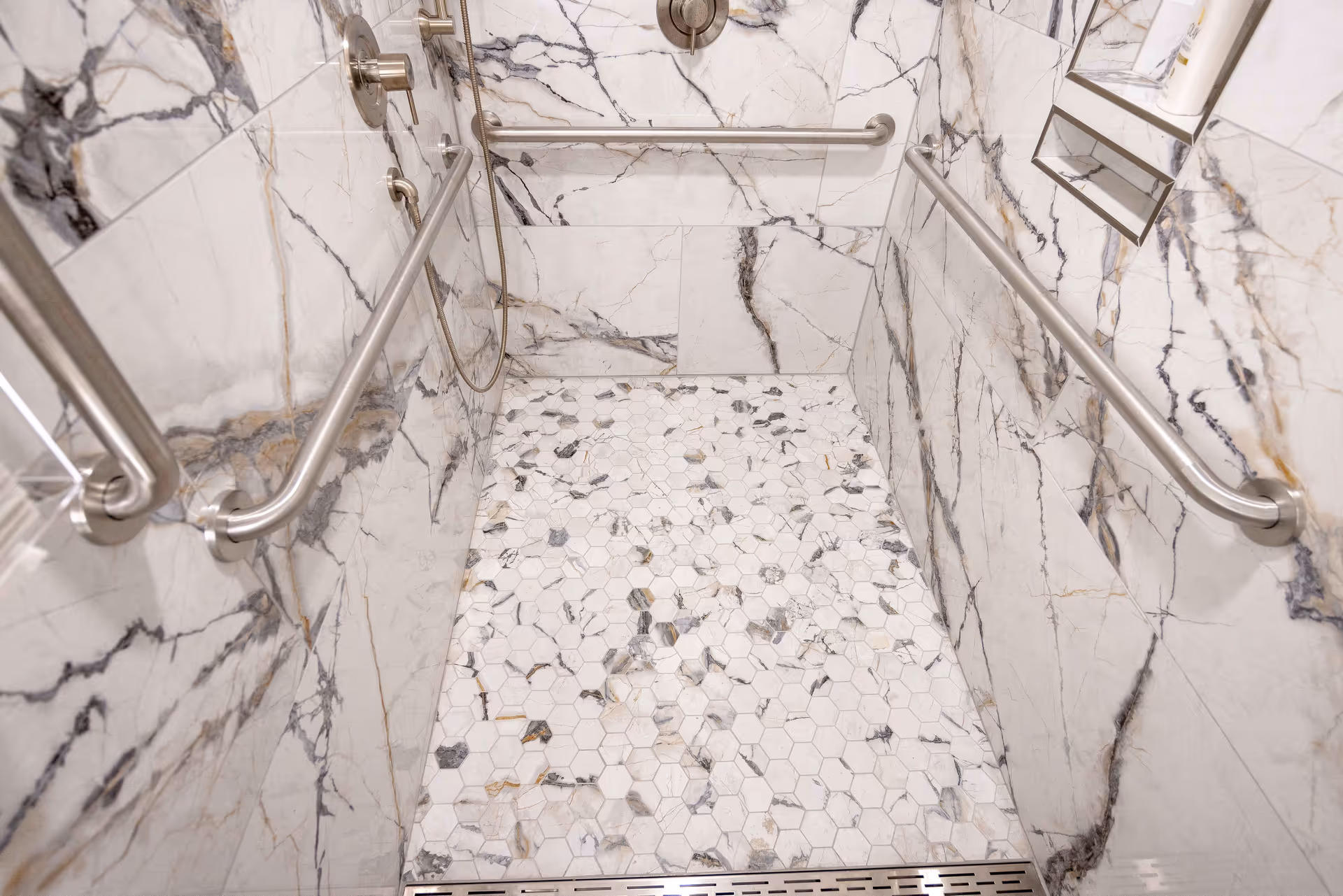 Wheelchair-accessible marble-tiled shower stall with hexagon mosaic floor, stainless steel grab bars, and a handheld shower head.