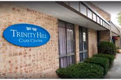 Exterior view of a brick building with a blue oval sign that reads 'Trinity Hill Care Center'. There are windows with curtains and neatly trimmed green bushes along the sidewalk.