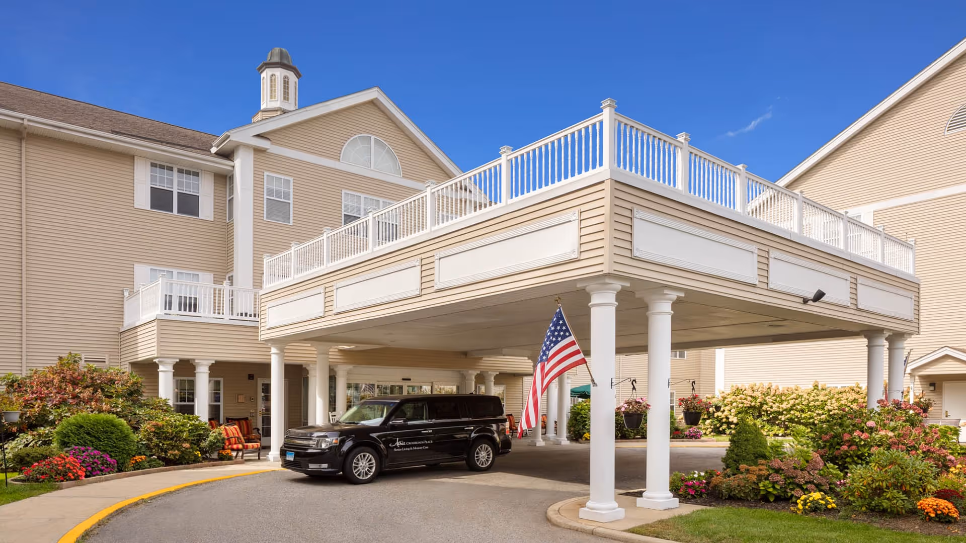 Exterior view of Atria Crossroads Place senior living facility showing a covered driveway with white columns, a black vehicle parked under the canopy, an American flag, and well-maintained landscaping with colorful flowers and shrubs under a clear blue sky.
