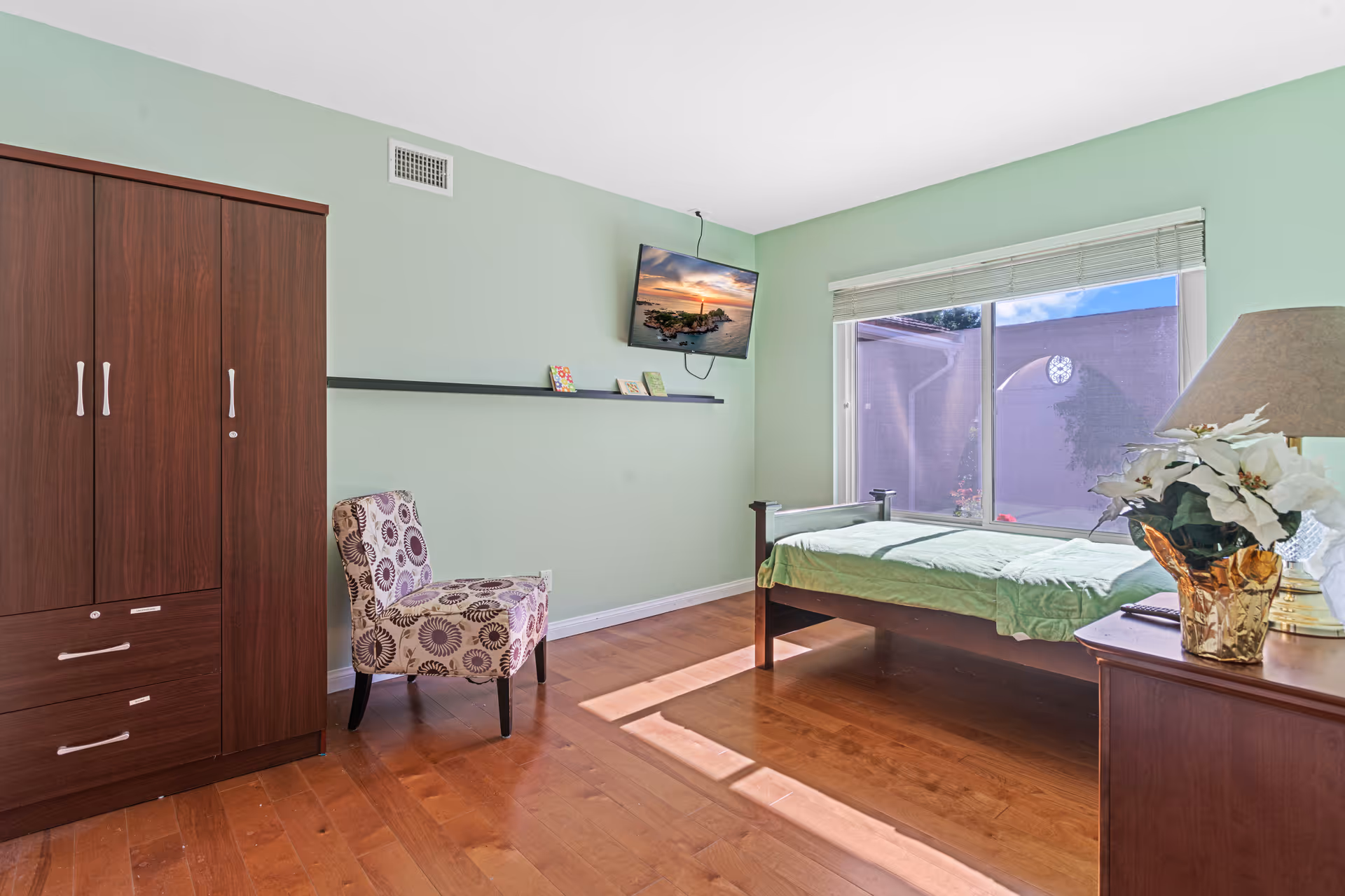 A bright bedroom with light green walls and wooden flooring. The room features a single bed with a green bedspread positioned near a large window with blinds, allowing natural light to fill the space. There is a wooden wardrobe on the left side, a patterned upholstered chair next to it, and a wooden nightstand with a lamp and a vase of white flowers on the right. A flat-screen TV is mounted on the wall above a narrow shelf holding small decorative items.