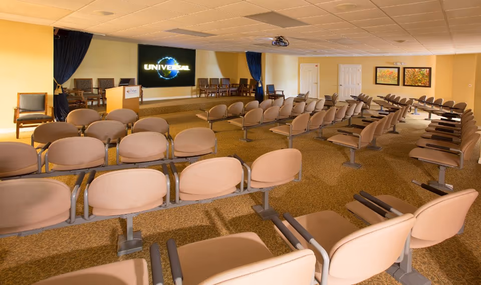 A large room set up as a presentation or meeting space with multiple rows of beige chairs facing a stage with a podium and a large screen displaying the Universal logo. The room has yellow walls, carpeted floor, and framed artwork on the far wall.