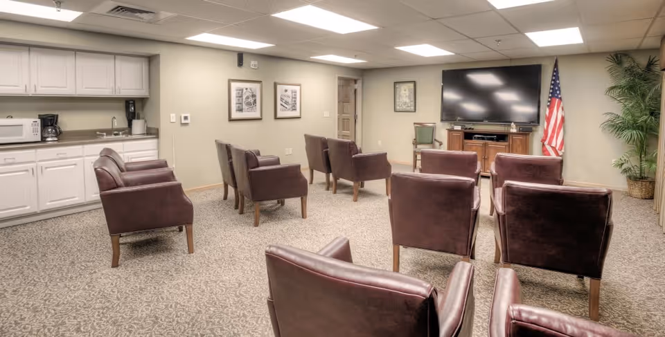 A common area with several brown leather chairs arranged in rows facing a large flat-screen TV mounted on a wooden cabinet. An American flag stands to the right of the TV, and a large potted plant is in the corner. On the left side of the room, there is a kitchenette with white cabinets, a microwave, a coffee maker, and a sink. The walls are beige, and there are framed pictures hanging on the wall.