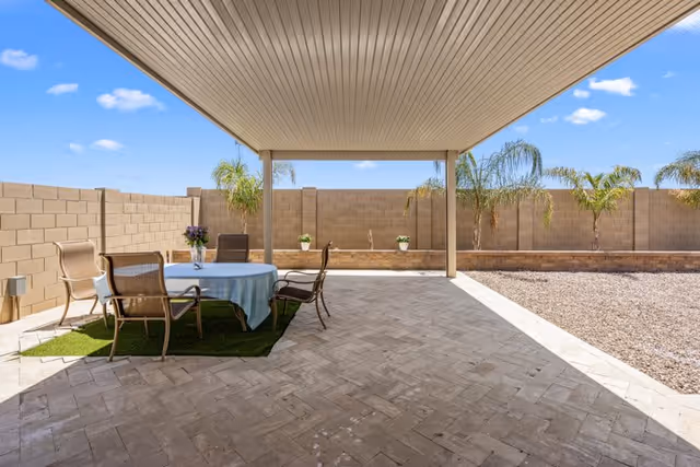 Covered outdoor patio area with a round table covered by a light blue tablecloth and surrounded by four brown chairs. The patio is paved with light-colored tiles and has a small patch of artificial grass under the table. In the background, there is a beige brick wall with several small palm trees and potted plants. The sky is clear and blue.