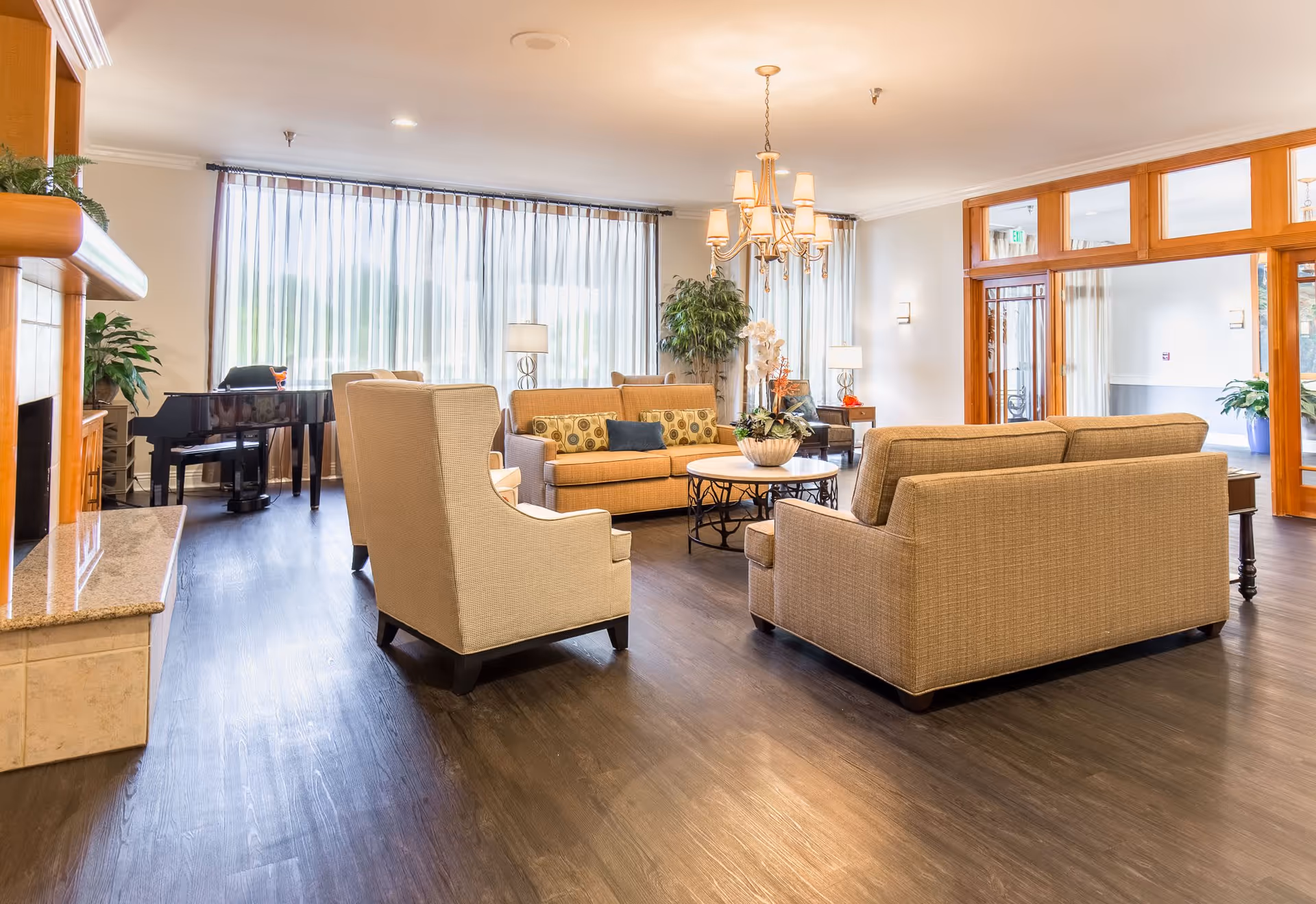 A bright and spacious assisted living facility common area with beige sofas and armchairs arranged around a round coffee table with a floral centerpiece. Large windows with sheer curtains allow natural light to fill the room. A grand piano is positioned near the windows, and a chandelier hangs from the ceiling. The room features wooden flooring and light-colored walls with wooden trim around the doors and windows.