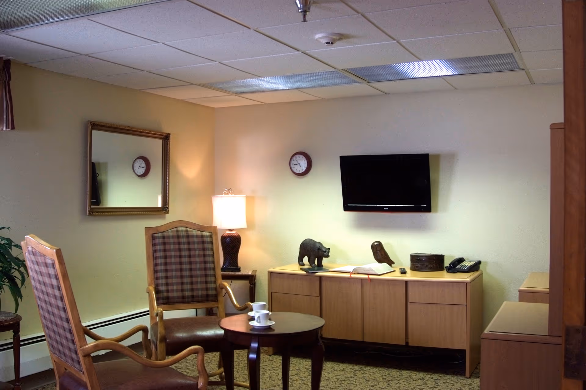 Cozy sitting room with two plaid-upholstered chairs around a small round table, a lamp, wall-mounted TV, and a low cabinet with decorative items.
