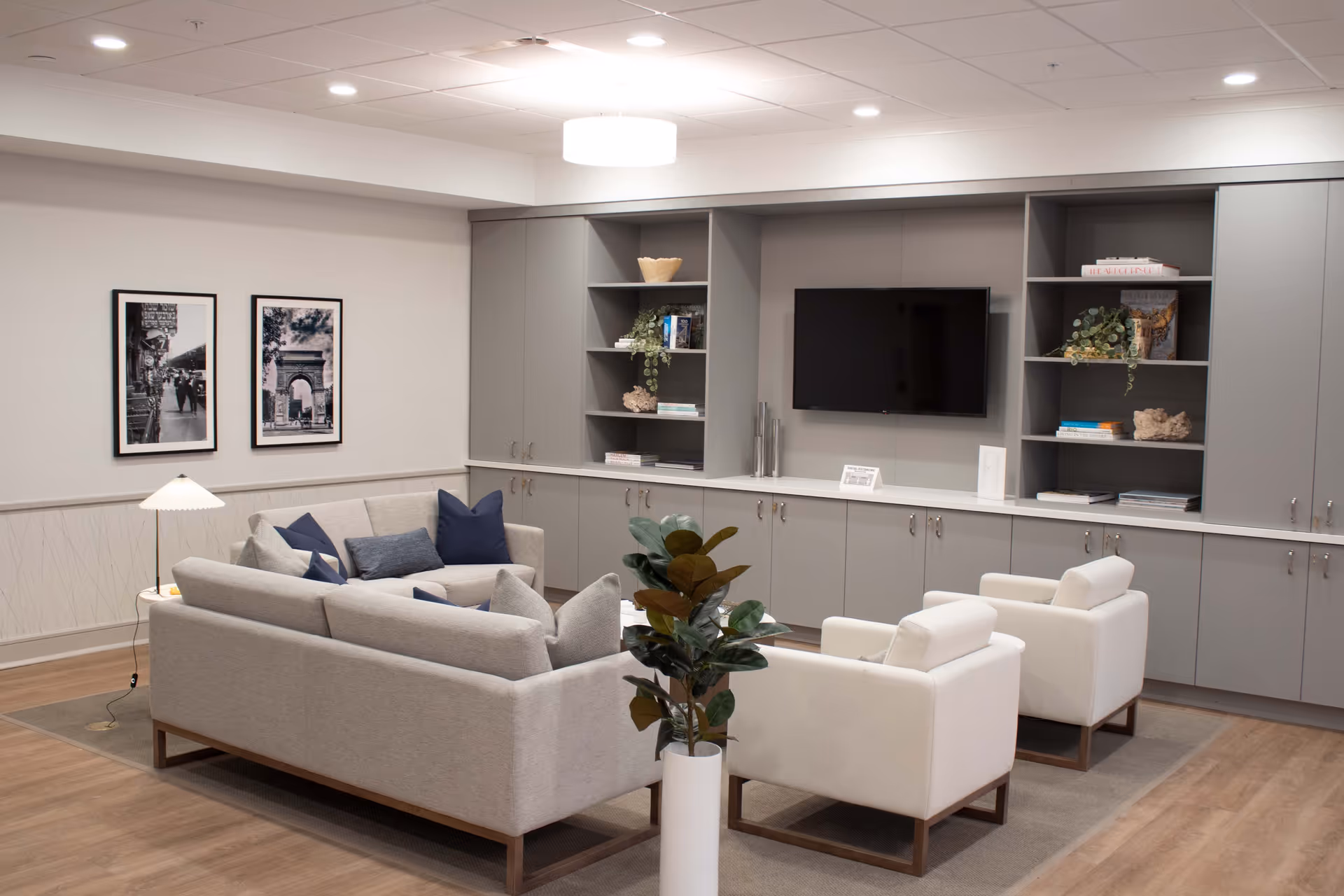 A modern living room area with a light gray sofa and two white armchairs arranged around a coffee table. Behind the seating is a built-in gray shelving unit with cabinets, decorative items, books, and a wall-mounted flat-screen TV. The room has wood flooring, a floor lamp, two framed black and white photos on the wall, and a potted plant in the foreground.