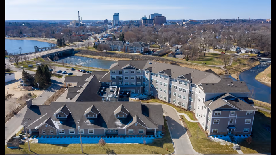 Aerial view of the AVIVA River Bend senior living complex beside a river with nearby roads and the town skyline in the background.