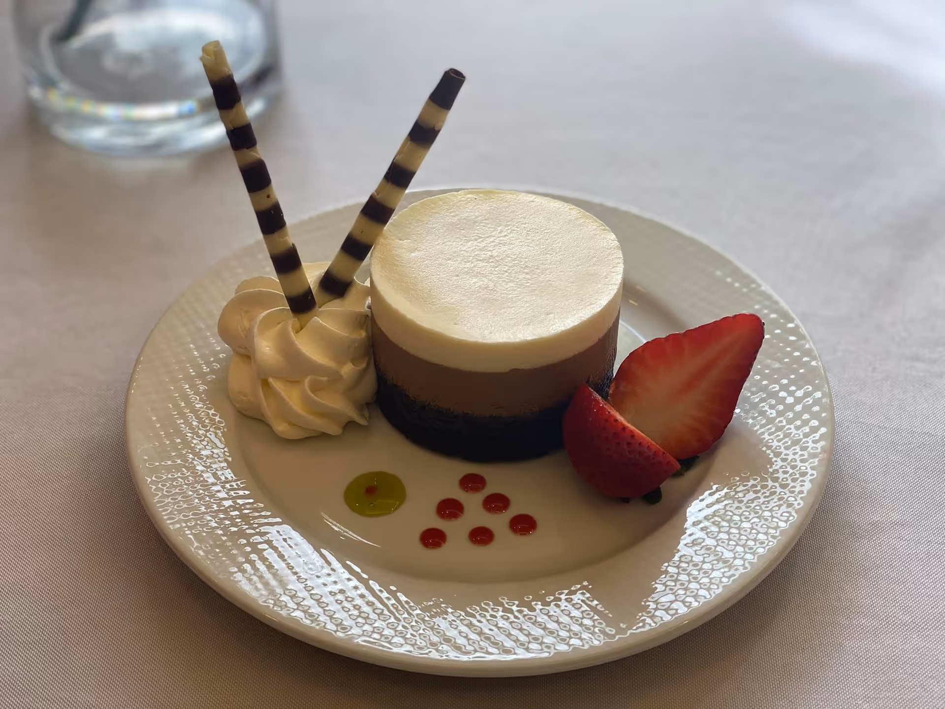 Plated layered mousse dessert topped with whipped cream, two striped chocolate wafer sticks and a halved strawberry on a white plate.